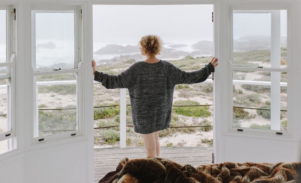 A woman stands in a beach house looking out to the water